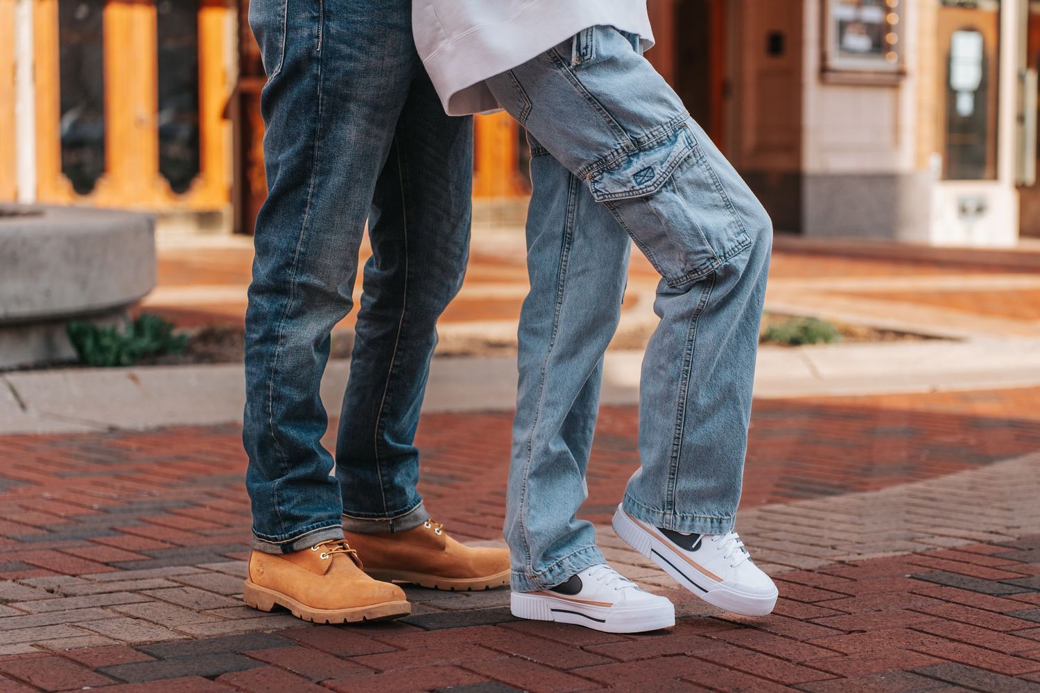 Close-up of two people's legs in jeans and contrasting footwear standing on brick pavement.
