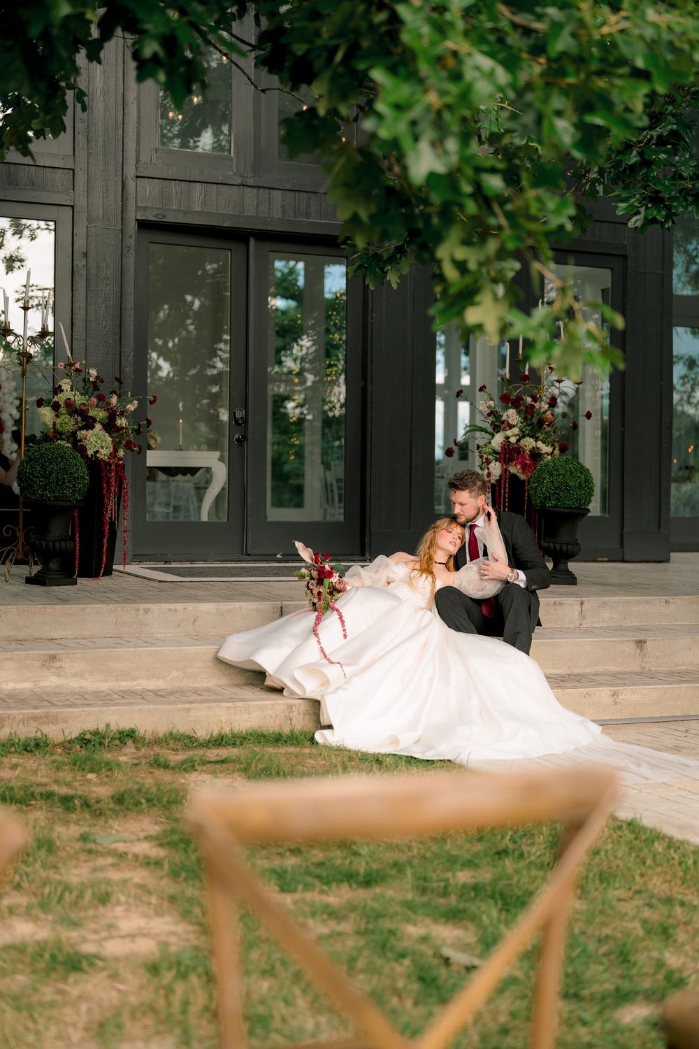 Bride and groom sitting on steps outside elegant dark building, bride in white gown holding burgundy bouquet.