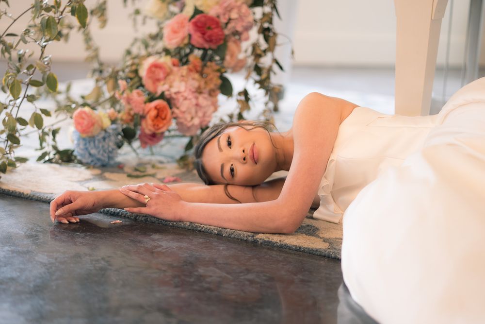 Bride in white gown lying gracefully on marble floor surrounded by lush pink and coral floral arrangements.