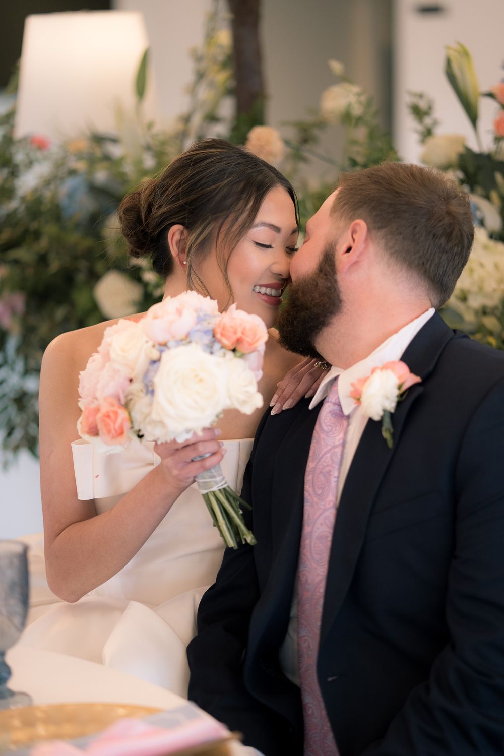 Bride and groom share a tender moment, she holds a pink and white bouquet, he wears a dark suit with pink tie.