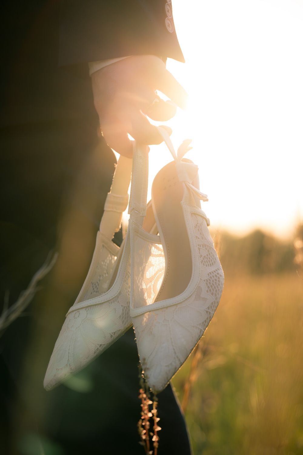 Hand holding white lace bridal heels against golden sunset light in a field.