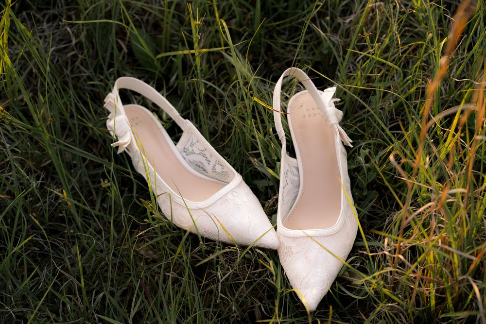 White lace slingback bridal heels with bow detail resting on green grass.