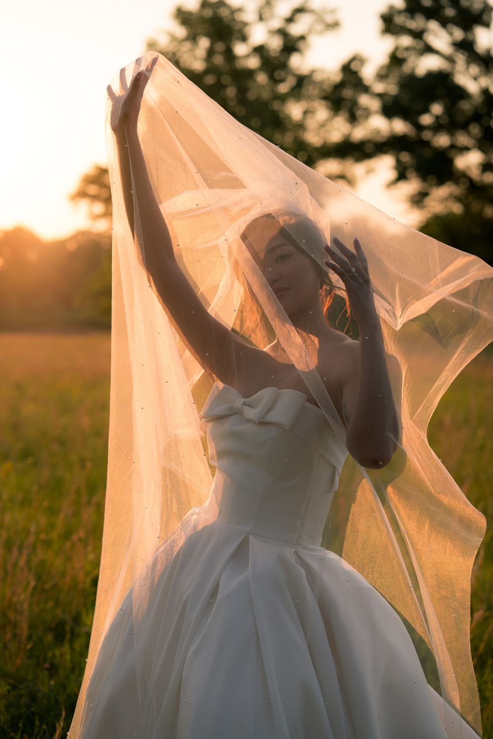 Bride in white ballgown lifting her veil against a golden sunset in an open field.
