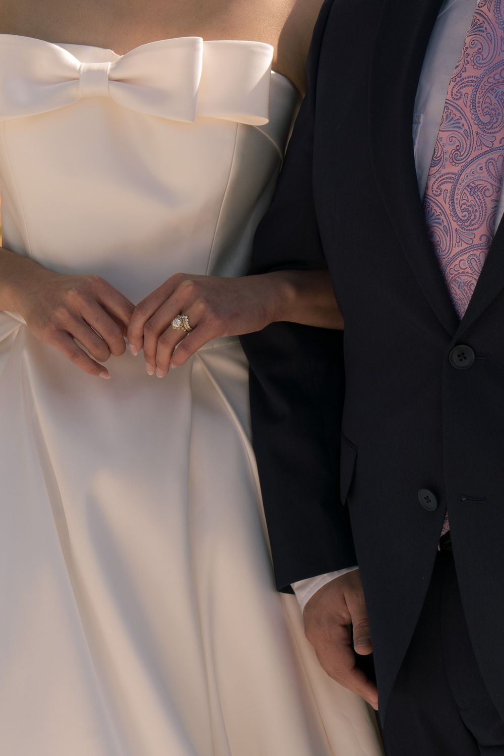 Close-up of bride and groom holding hands, showcasing wedding ring, white bow dress and dark suit with paisley tie.