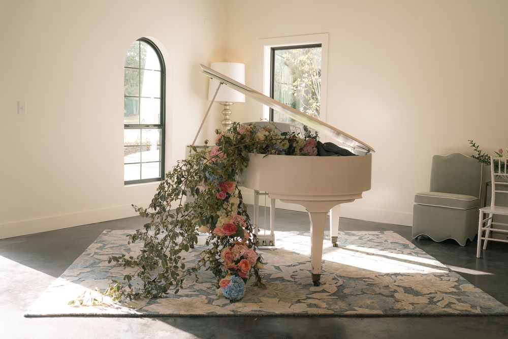 Elegant white baby grand piano adorned with cascading floral greenery and pink roses in a sunlit wedding venue.