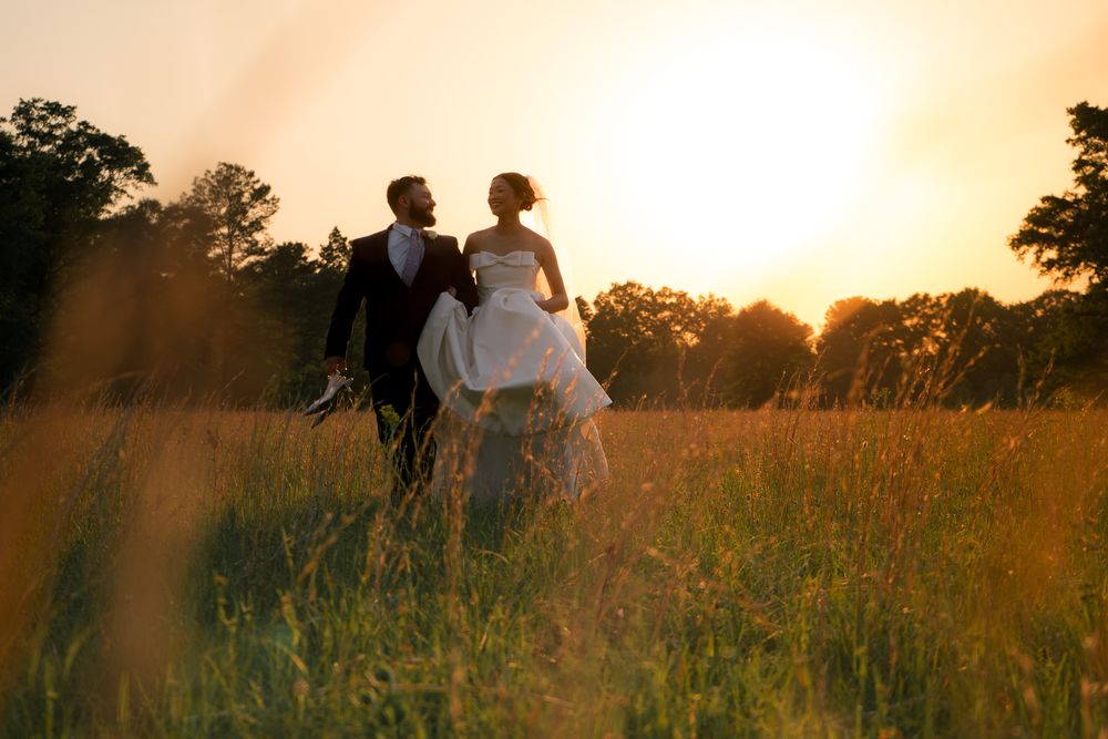 Bride and groom walking through golden meadow at sunset, bride holding dress, trees silhouetted in background.