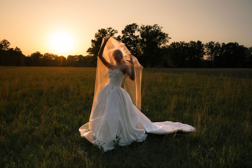 Bride in white gown holds flowing veil in sunlit field at golden hour sunset.