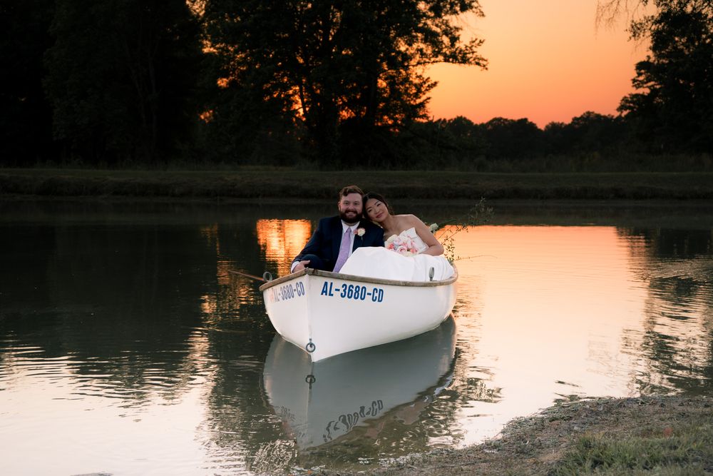 Newlyweds sitting in white rowboat on calm lake at sunset, sharing a romantic moment on their wedding day.