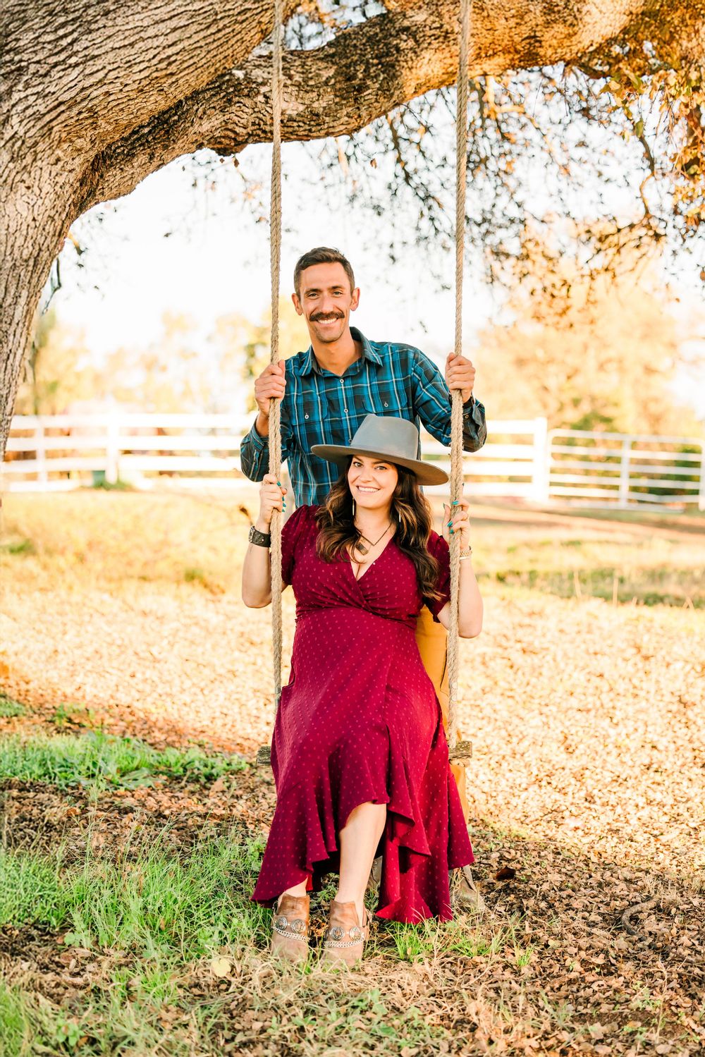 A couple sits together on a rustic tree swing in autumn with golden sunlight filtering through branches.
