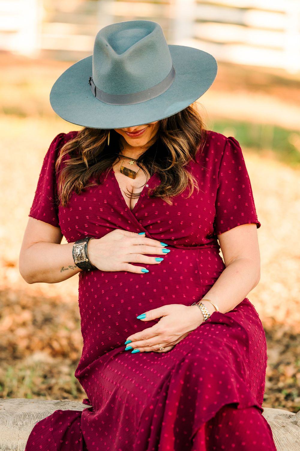 A pregnant woman in a burgundy polka dot dress and blue hat sits outdoors during golden hour.