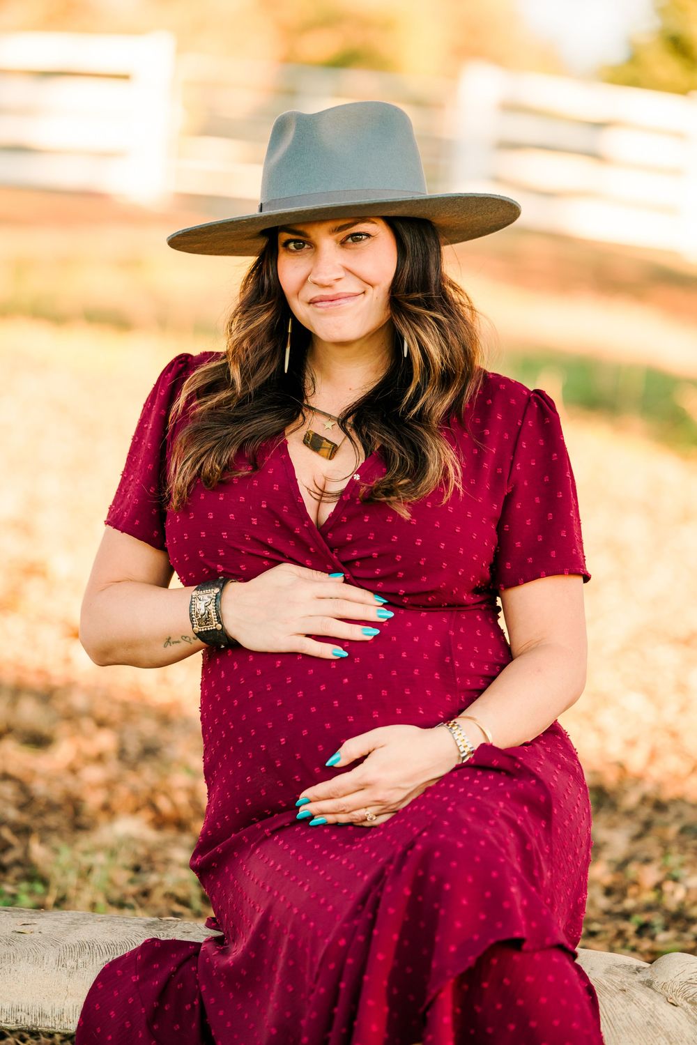 Pregnant woman in burgundy dress and wide brim hat sits outdoors during golden hour photography session.