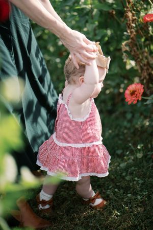 Baby wearing red gingham dress takes first steps on grass surrounded by flowers in garden setting.