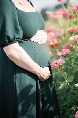 A pregnant person in an emerald green dress stands beside blooming pink flowers in a garden.