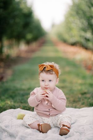A baby wearing a pink sweater and orange headband sits on a blanket between rows of apple trees.