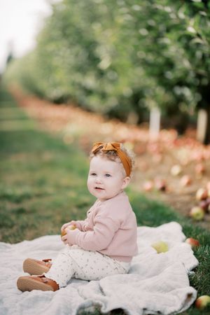 Baby in pink sweater and orange bow sitting on blanket during fall apple orchard photo session.