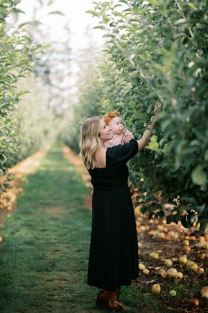 A figure in a long black dress reaches up to pick apples in a sunlit orchard.