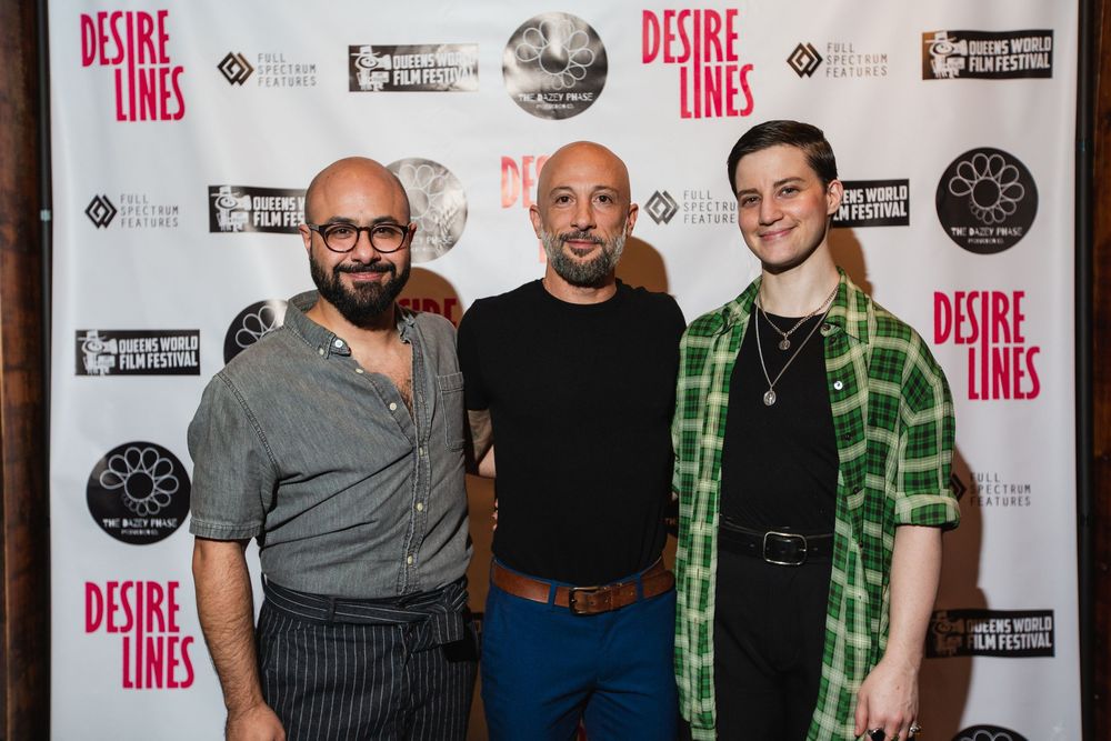 Three people posing against a promotional backdrop for 'Desire Lines' at an event.