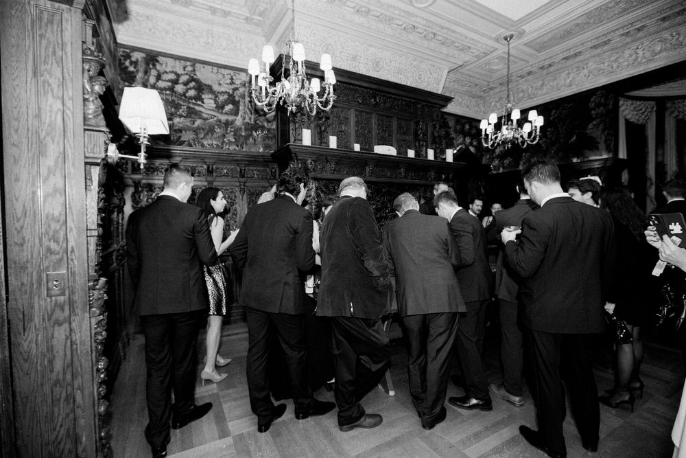 Formal event guests mingling in an elegant wood-paneled room with chandeliers.