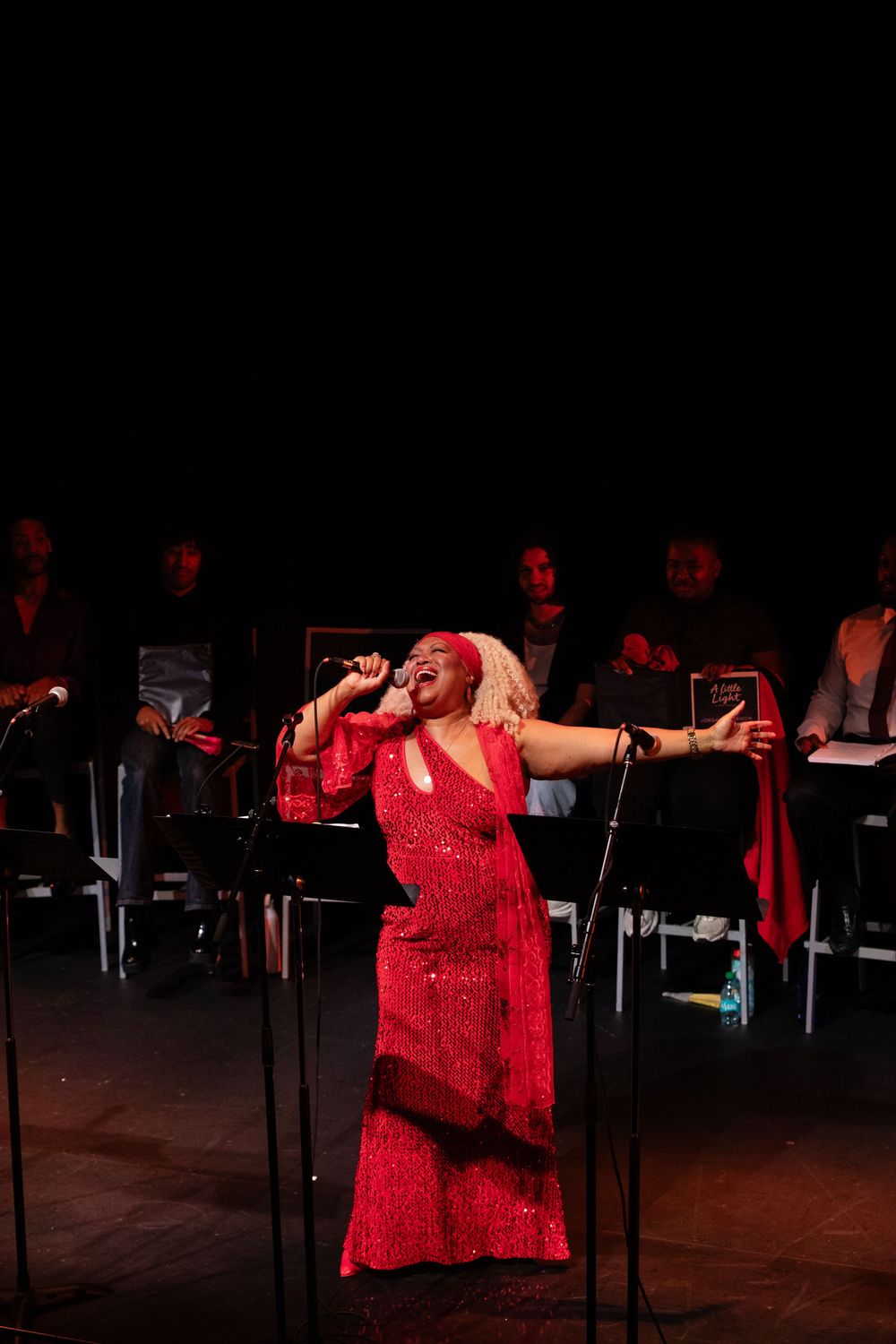 Woman in red sequin gown singing passionately on stage with musicians seated behind her in dim lighting.