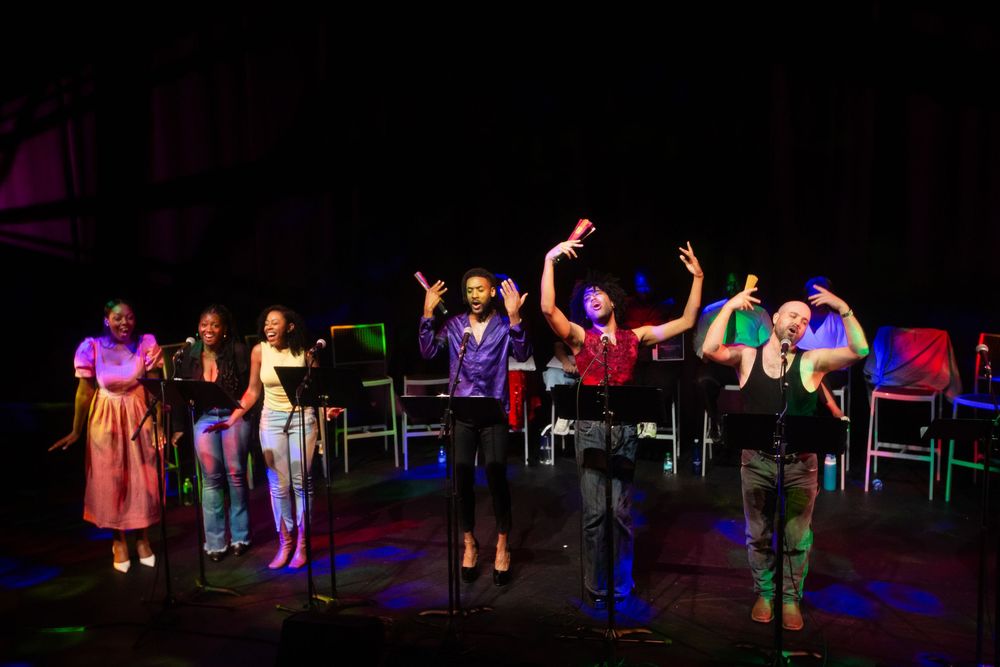 Diverse group of performers singing on a darkly lit stage with colorful lighting and microphone stands.
