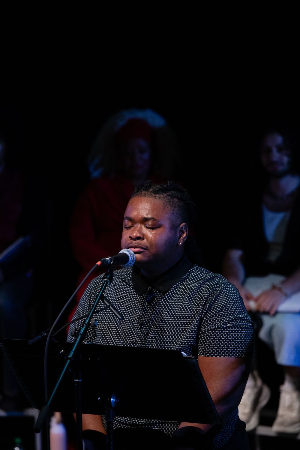 A man speaks into a microphone at a podium during a dimly lit stage performance with audience members seated behind him.