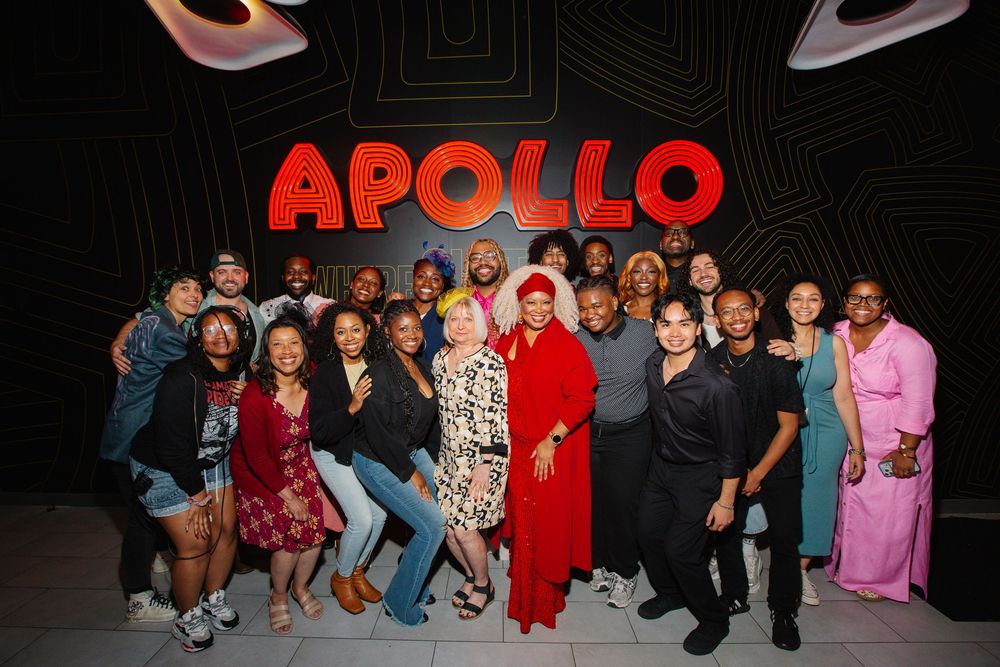 Diverse group of people posing together in front of illuminated Apollo sign at a special event.