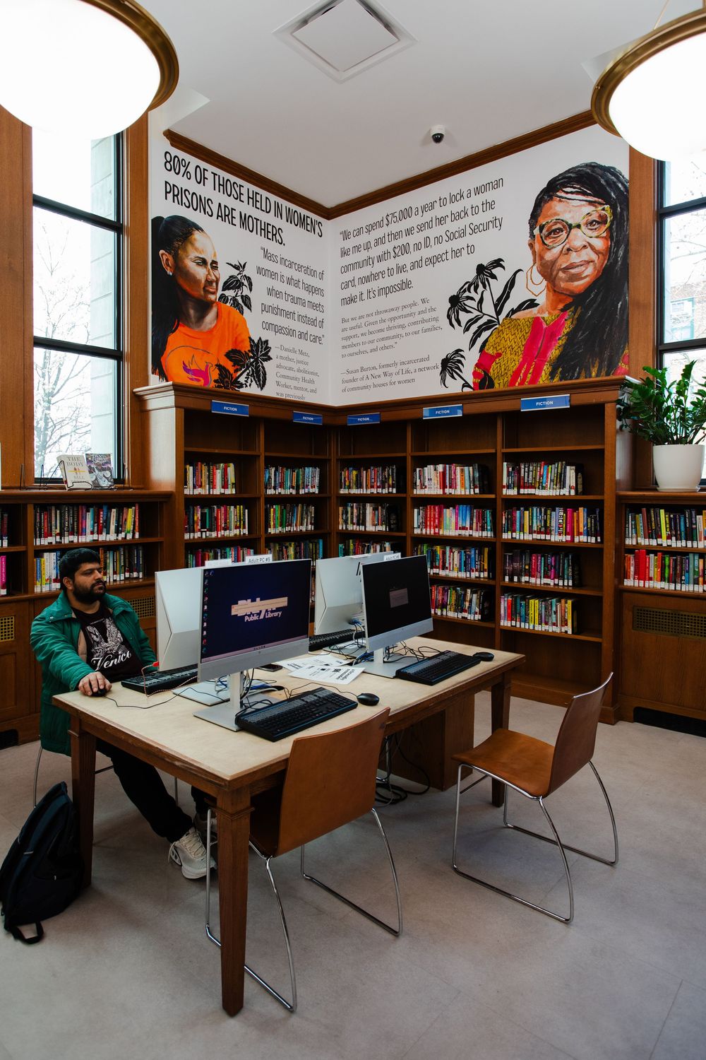 Library reading room with computer workstations, bookshelves, and a mural about women in prisons on the wall.