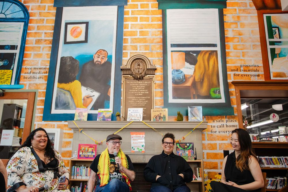 Four people sitting together in a colorful library with vibrant murals on brick walls behind them.