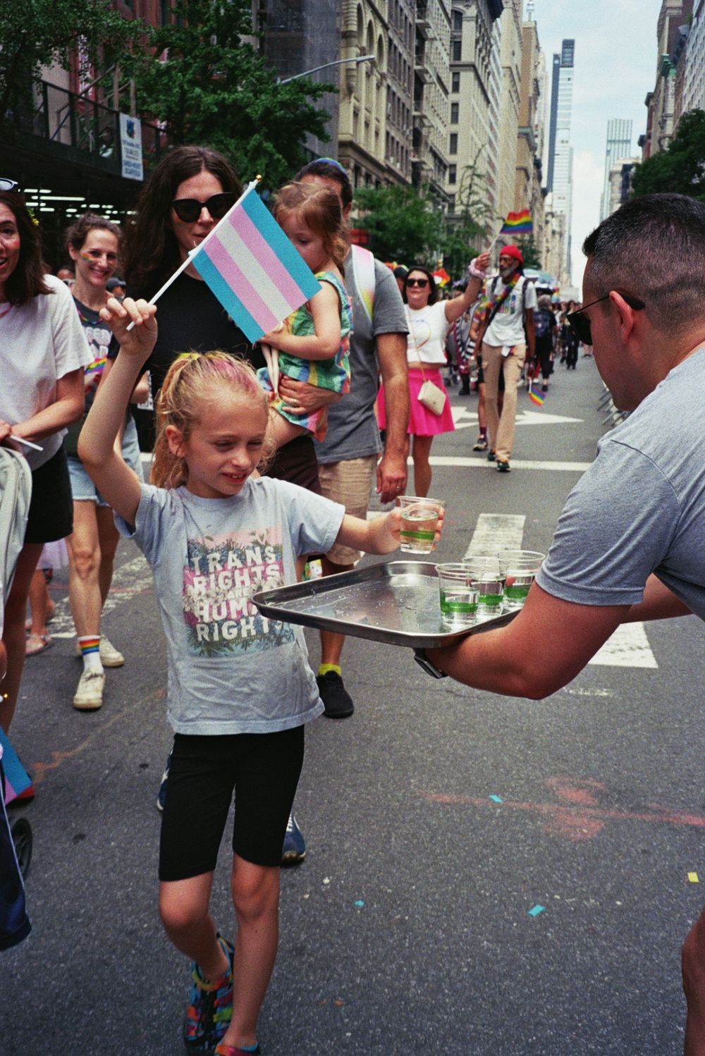 Young girl waves transgender pride flag while carrying drinks tray at NYC Pride parade, wearing Trans Rights Human Rights shirt.