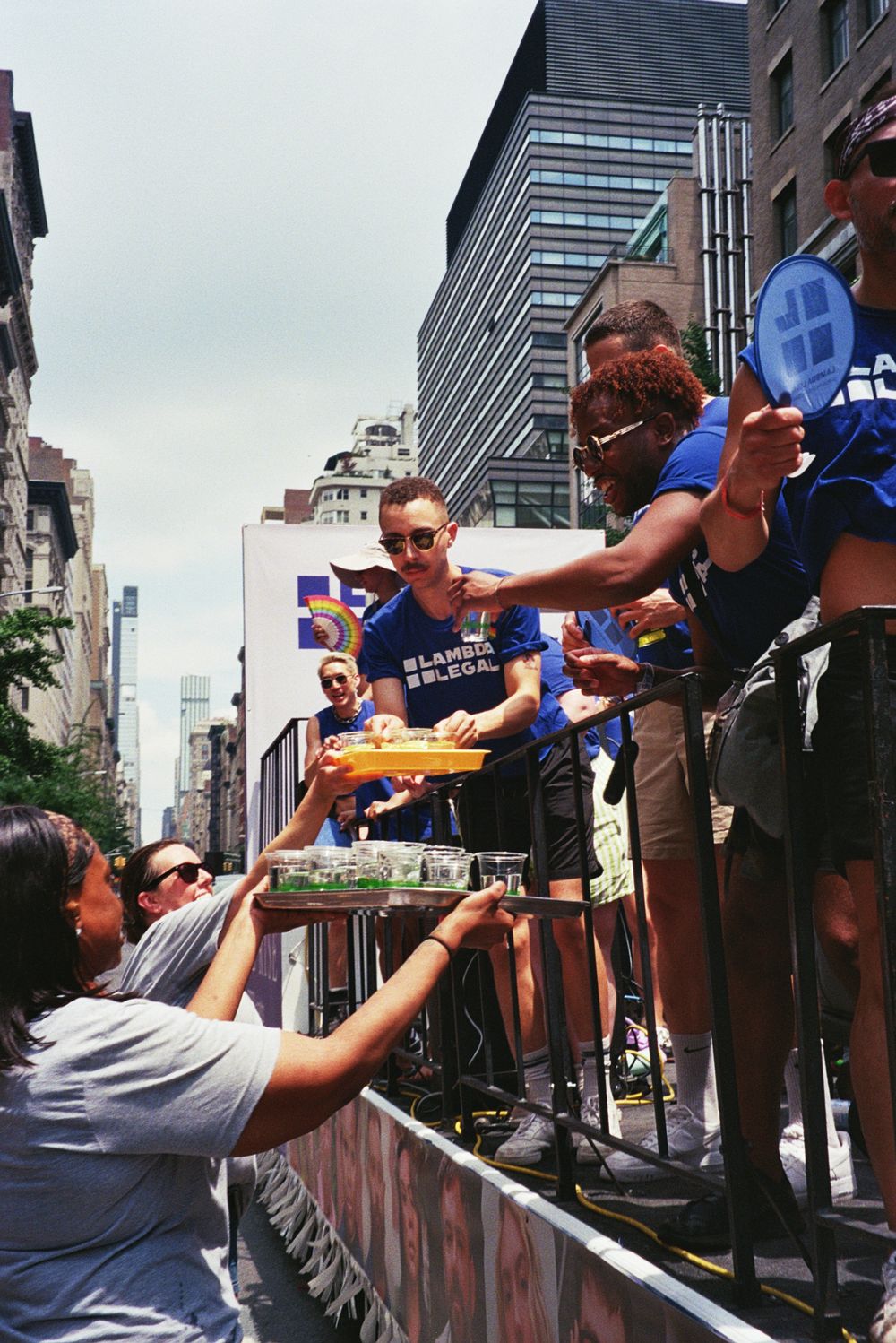 People handing drinks from a float to crowd during a sunny city parade on a bustling urban street.