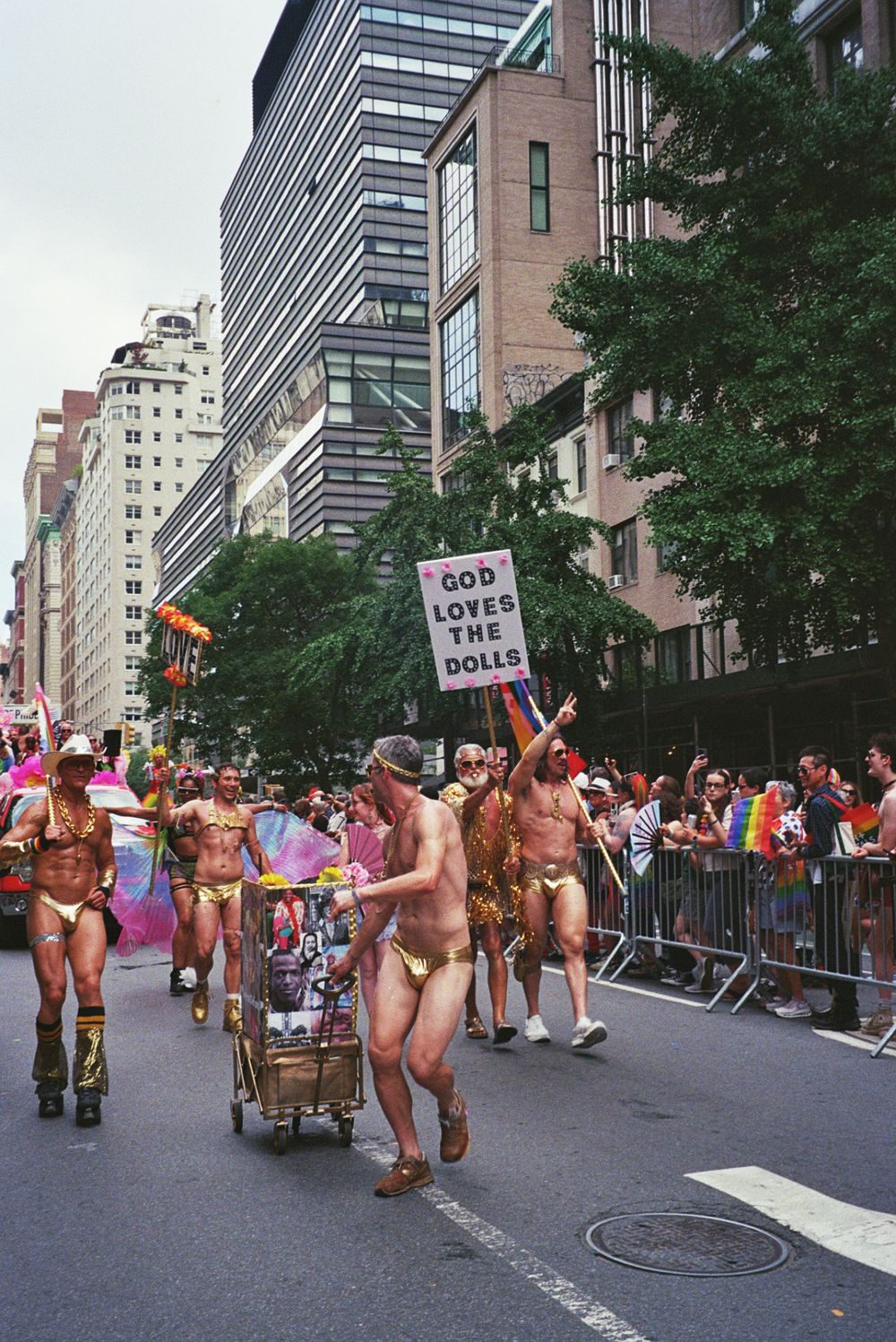 Participants in gold outfits march in a Pride parade on a city street, carrying a sign reading God Loves The Dolls.