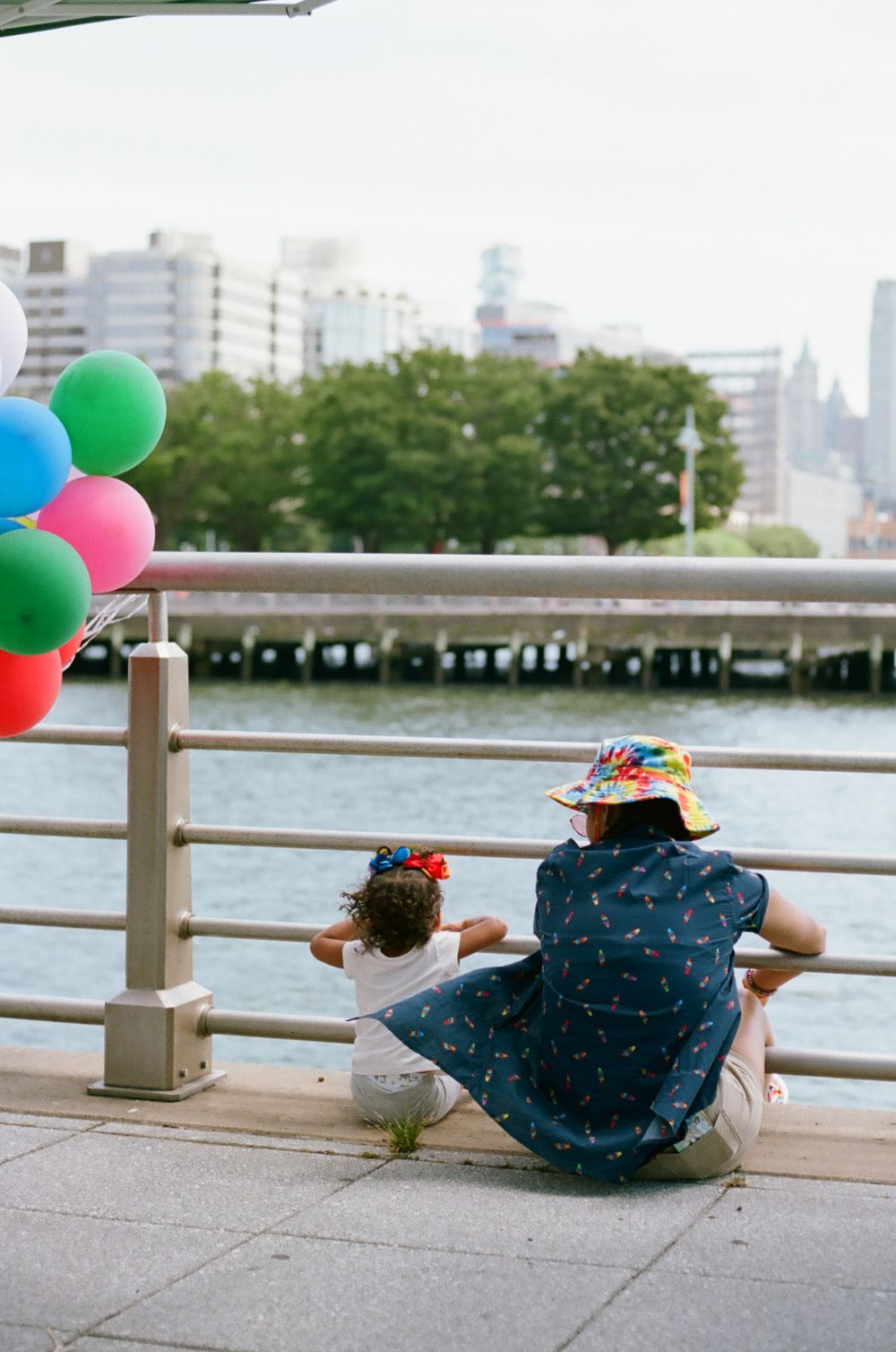 Parent and child sitting on waterfront deck with colorful balloons against city backdrop.
