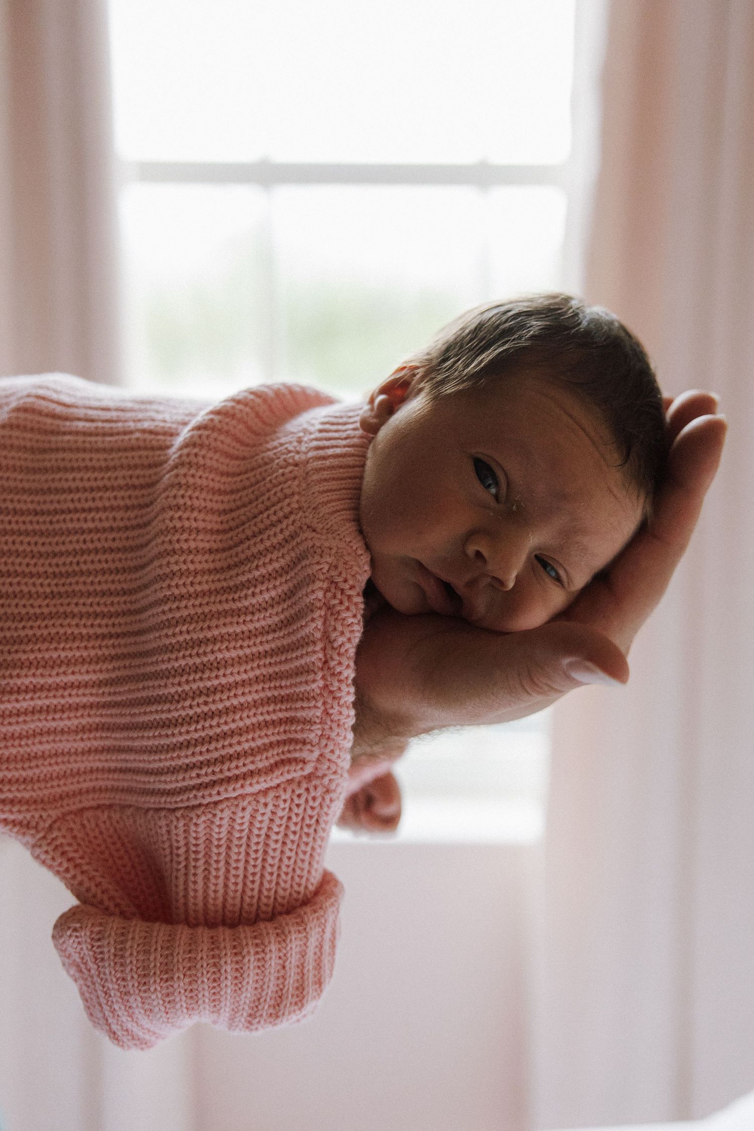 A small baby wrapped in a soft pink knit blanket gazes towards natural window light in a gentle indoor setting.