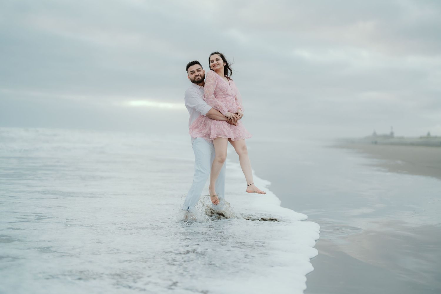 A romantic couple in pink outfits stands on an ice formation at a misty beach during wintertime.