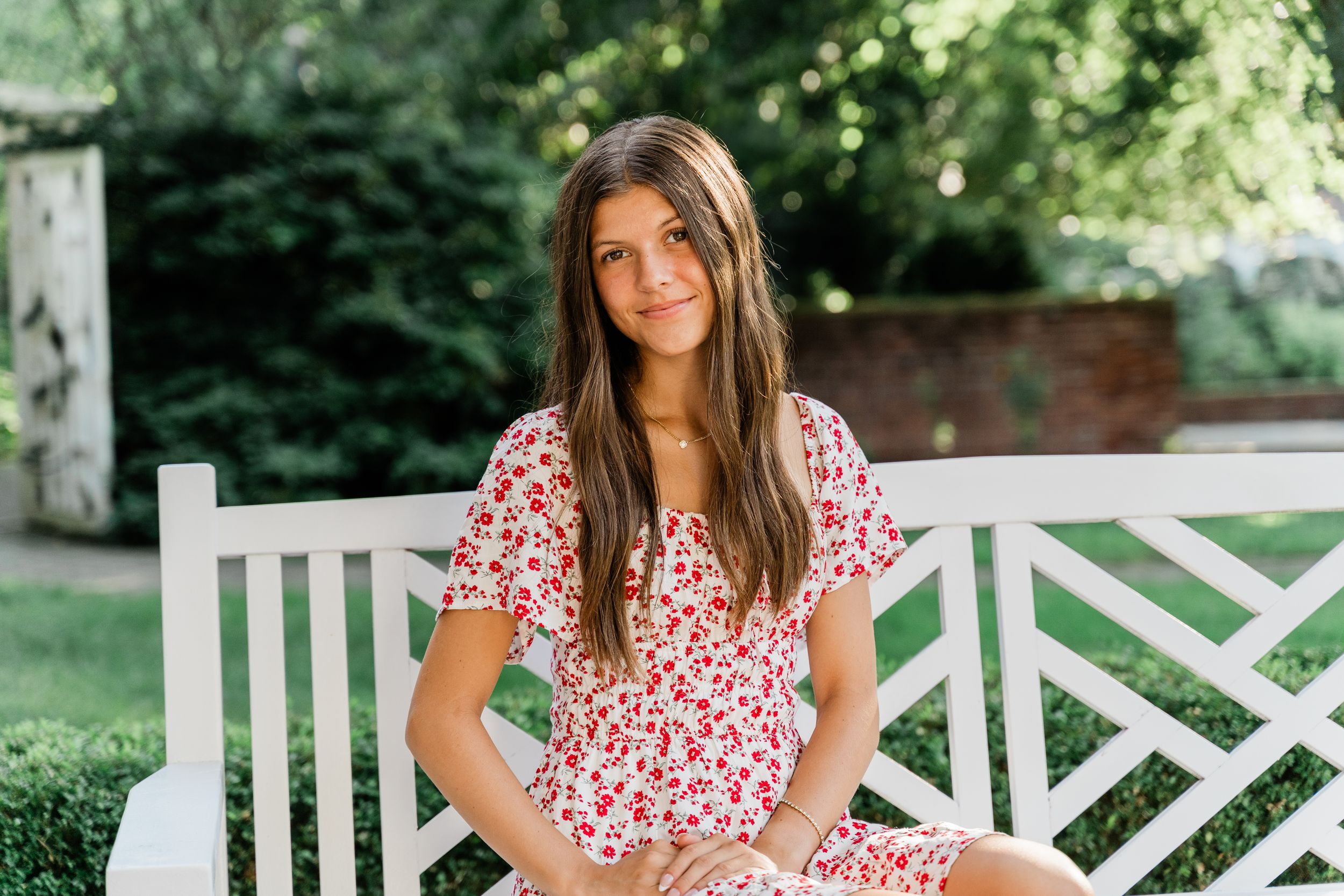 Woman in floral print dress sits on white wooden garden bench on sunny summer day.