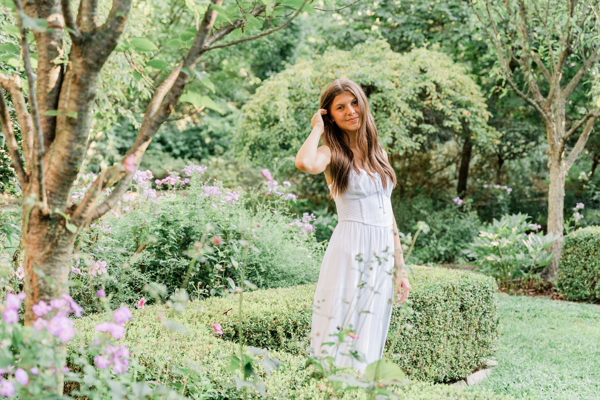 Someone in a white floral sundress poses in a lush summer garden with blooming flowers and manicured hedges.