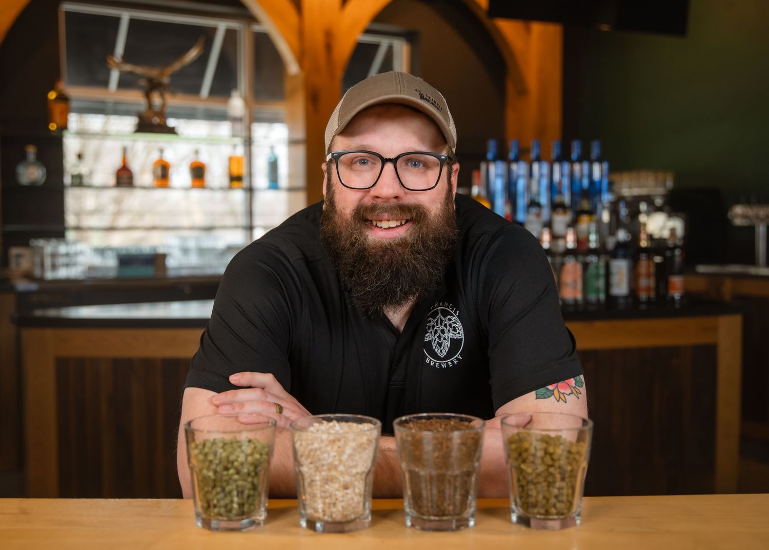 A bearded bartender shows multiple cocktail glasses with crushed ice and garnishes on a wooden bar counter.