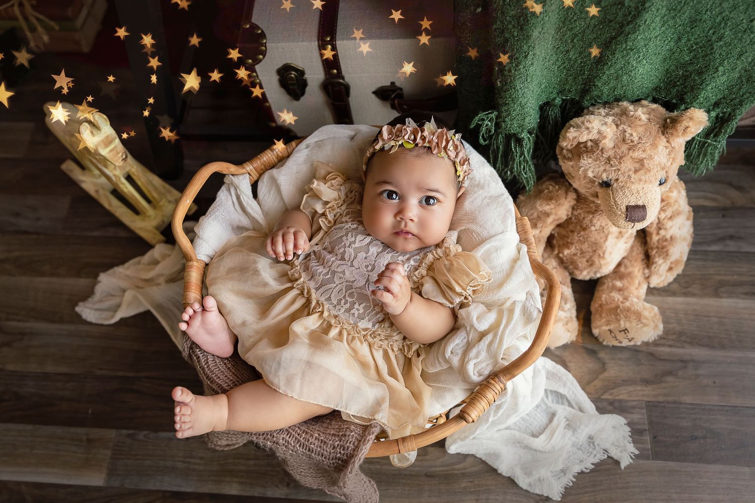 baby girl poses in a rattan basket looking up at the camera with teddy bear and rocking horse