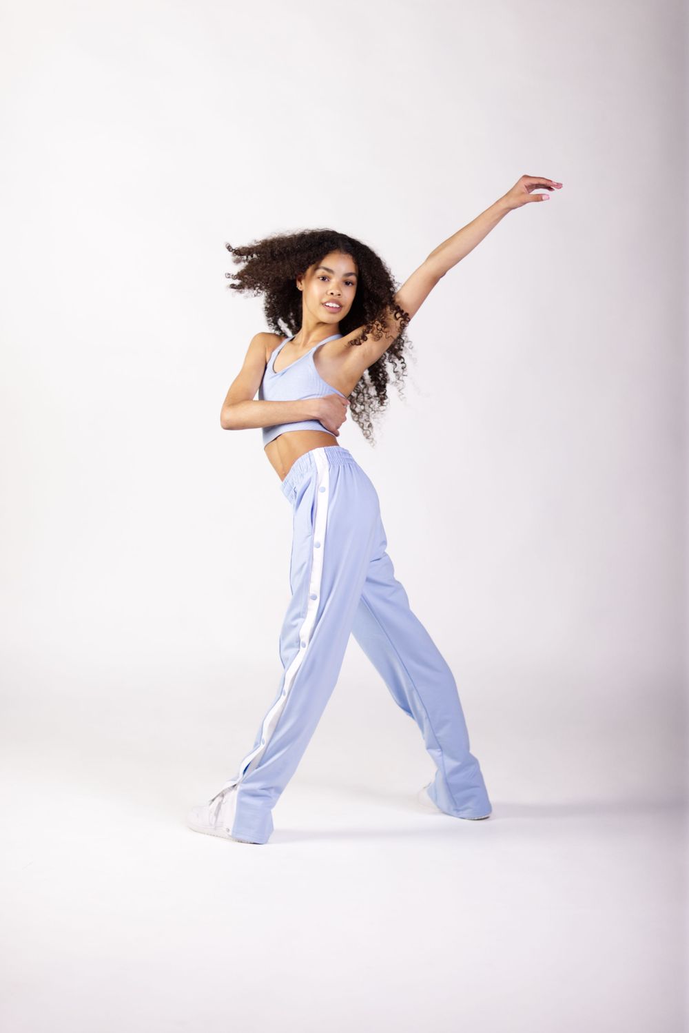 Dancer in light blue track pants and crop top strikes dynamic pose with flowing curly hair against white background.