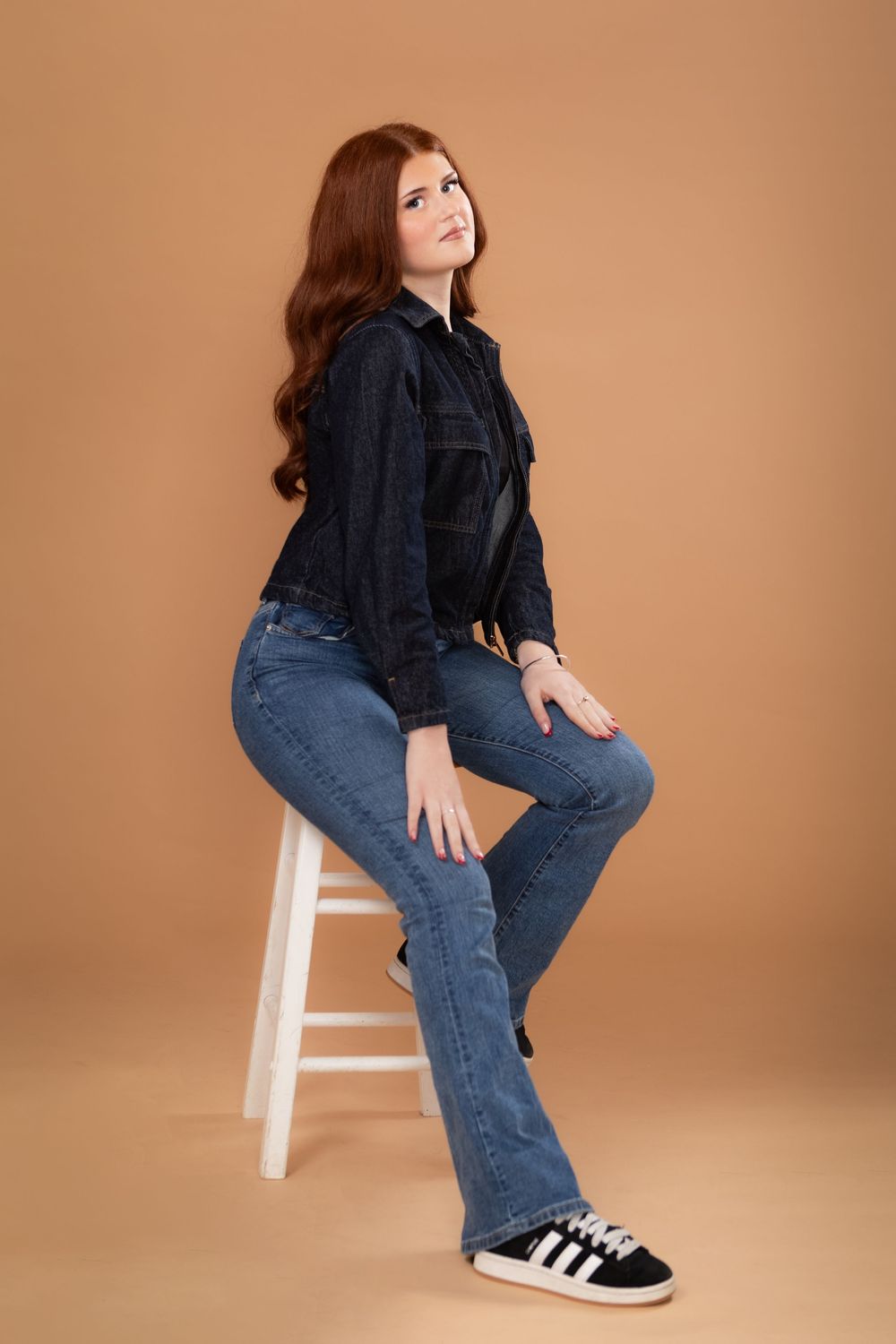Person in dark sweater and jeans sitting on white stool against brown background for fashion photography.