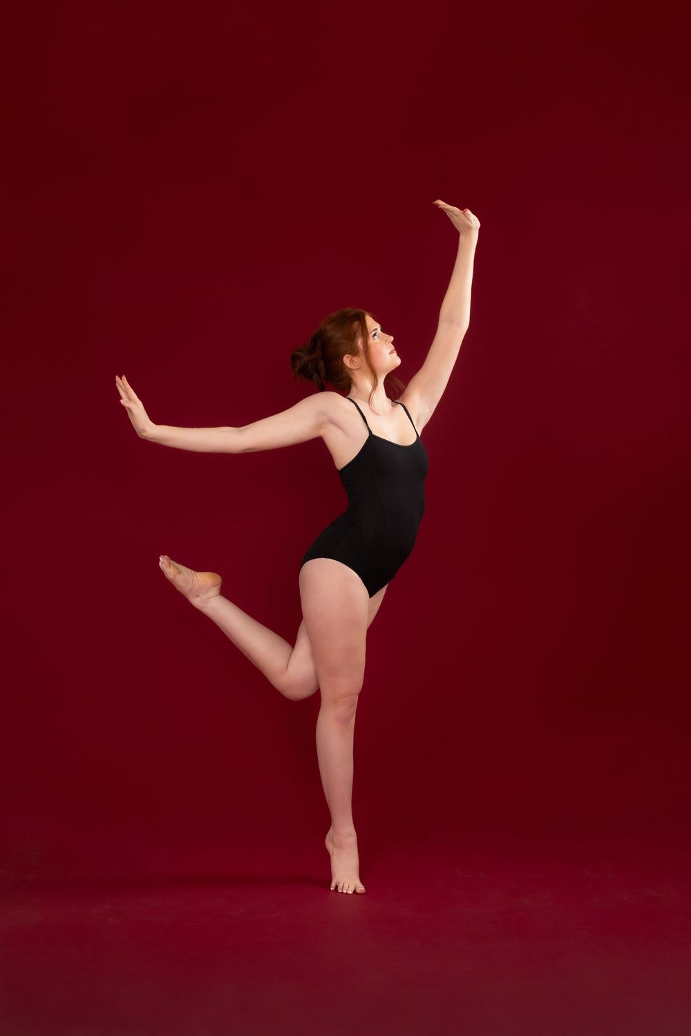 A ballet dancer in a black leotard performs a graceful arabesque pose against a deep burgundy background.