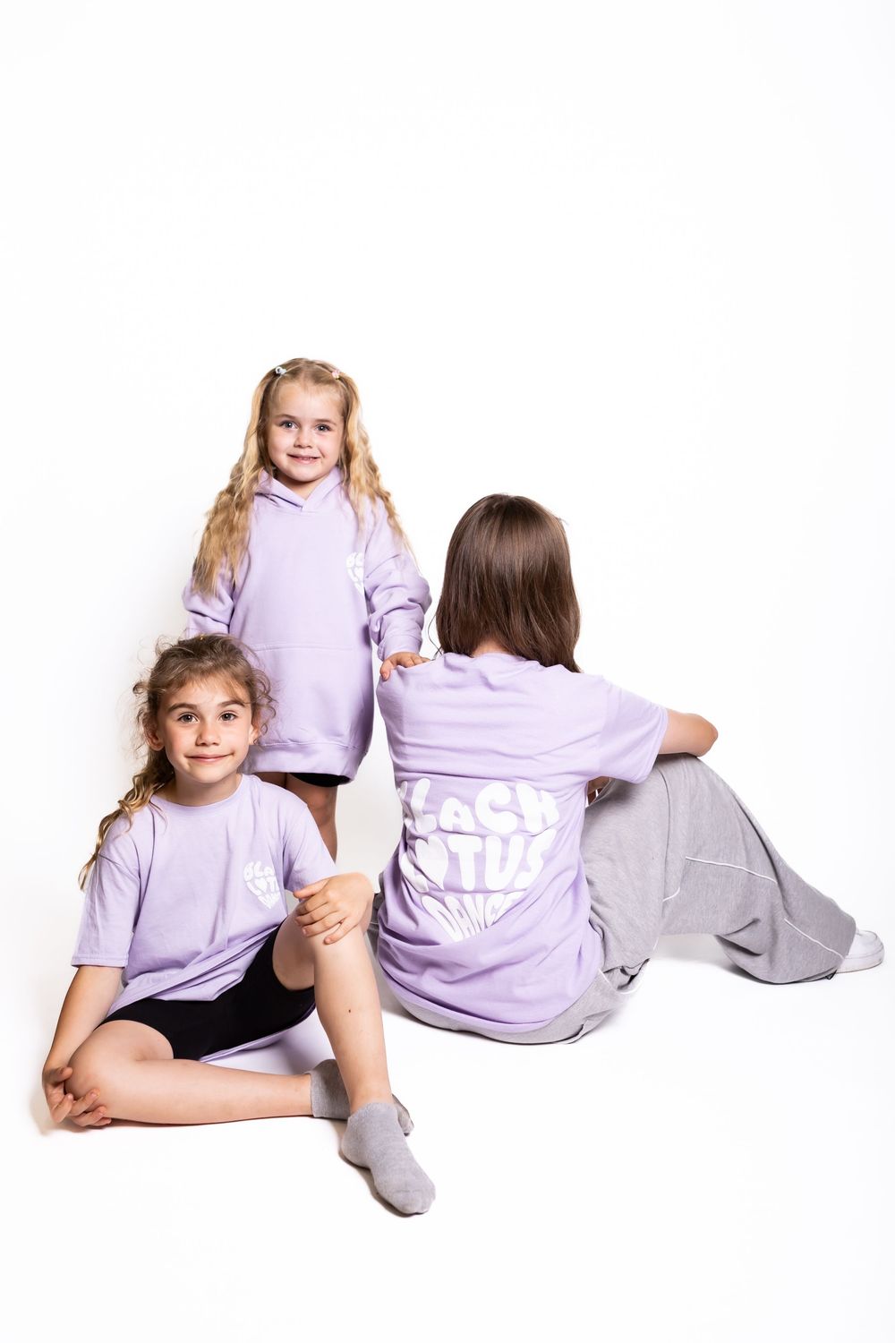 Three children wearing matching lavender colored tops and gray socks sitting and posing on white background.