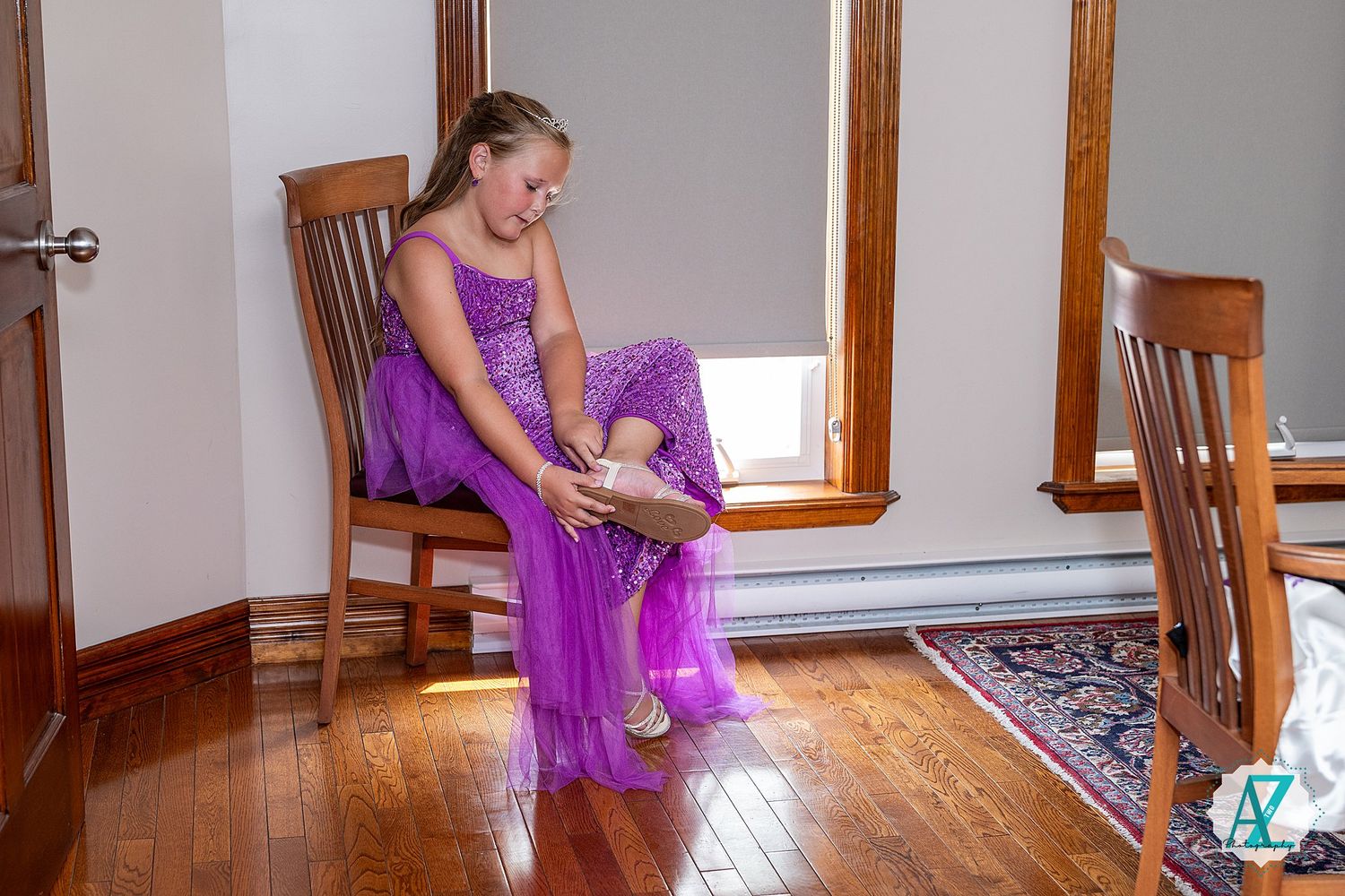 A person in a flowing purple dress sits on a wooden chair while putting on shoes.