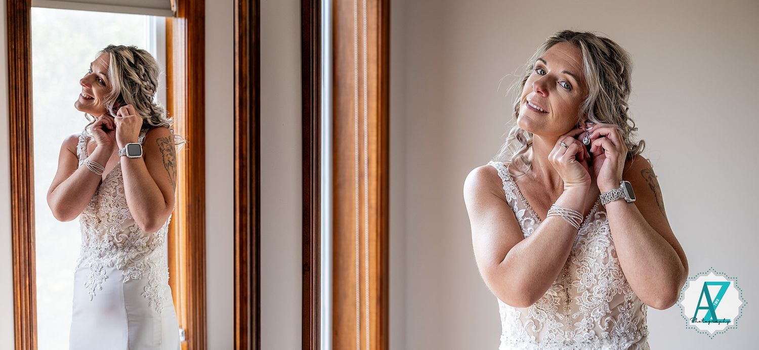 Bride in lace white wedding dress puts on earrings while standing near a wooden framed mirror.