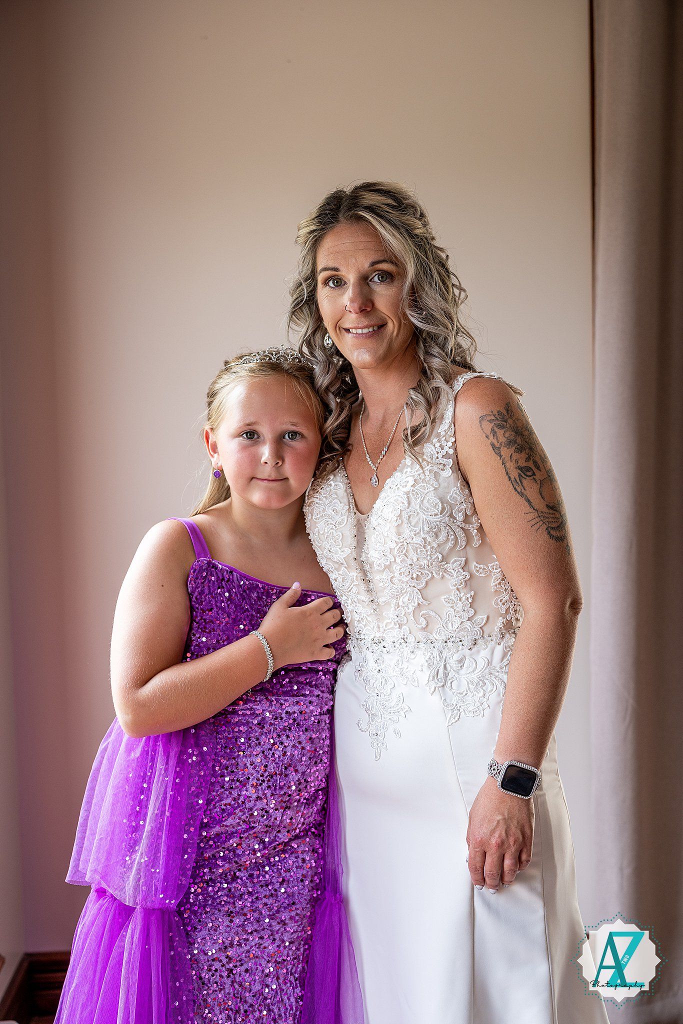 Two people pose together at a wedding, one wearing a white lace gown and the other in a sparkly purple dress.