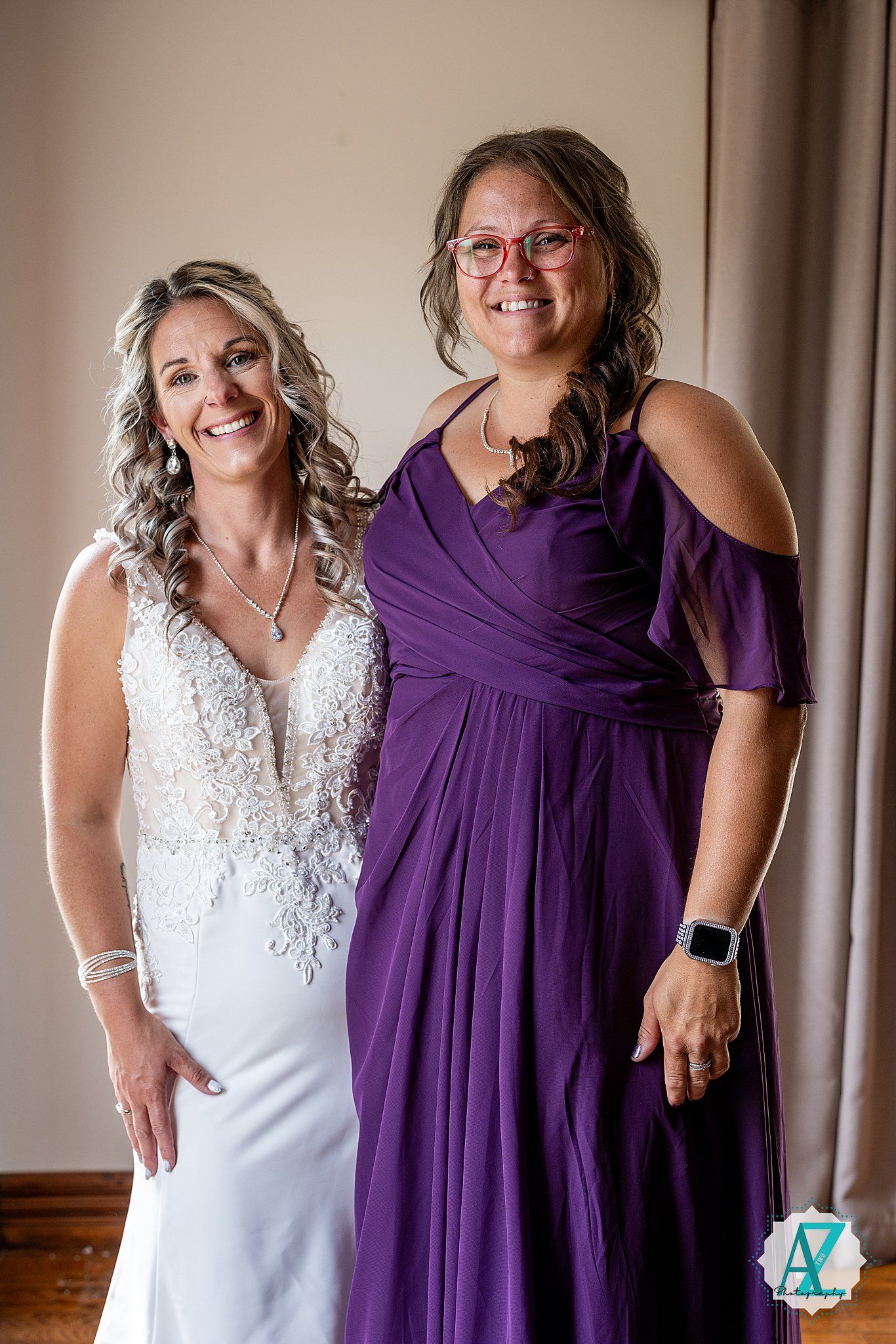 Two women stand close together at a wedding, one in a white bridal gown and one in a flowing purple dress.