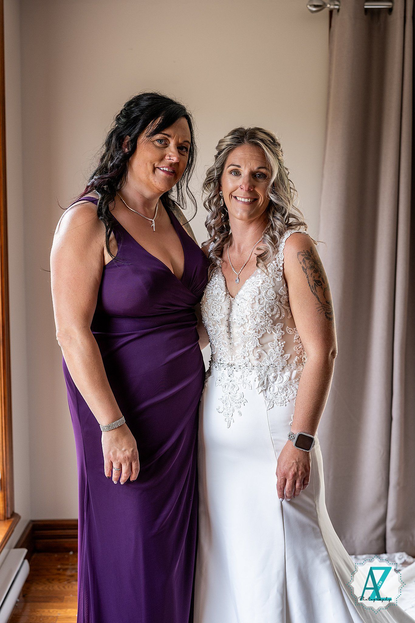 Two women pose together at a wedding celebration wearing a purple bridesmaid dress and white lace wedding gown.