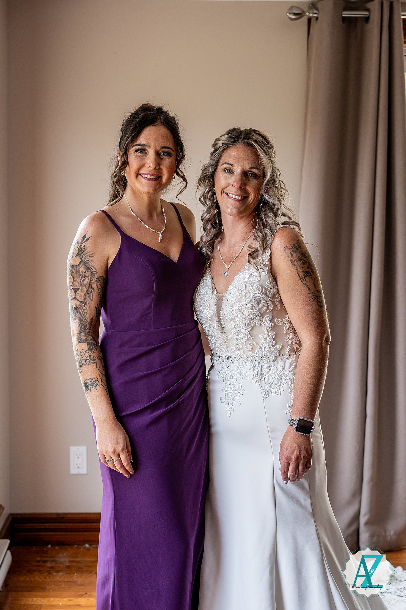 Two women in formal dresses, one wearing purple and one in white, pose together showing their arm tattoos.