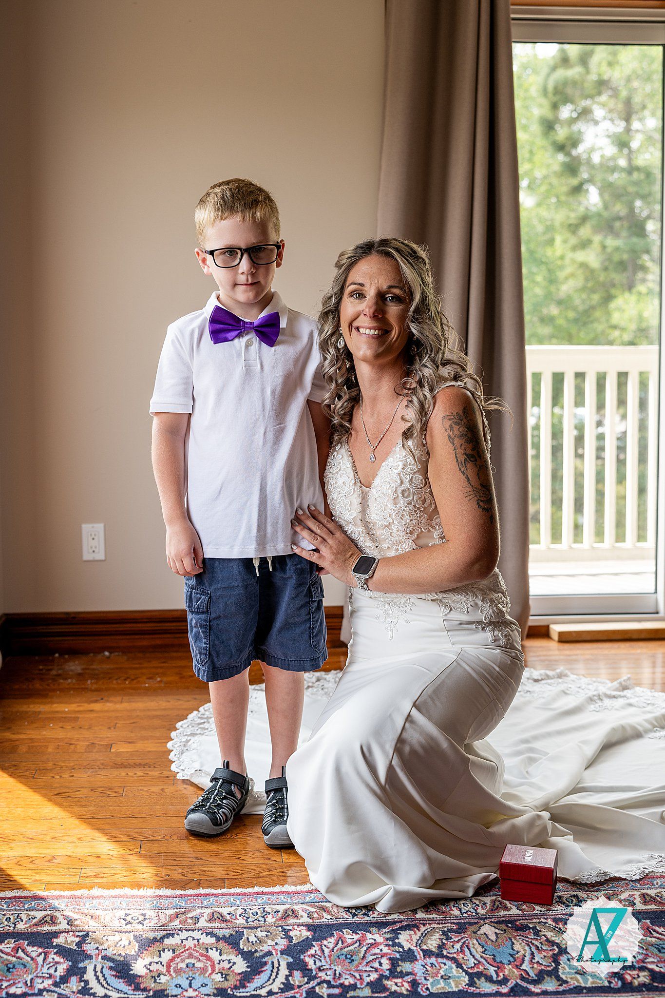 A formal picture of a bride in a white lace dress kneeling next to a young child in a bowtie on hardwood floors.
