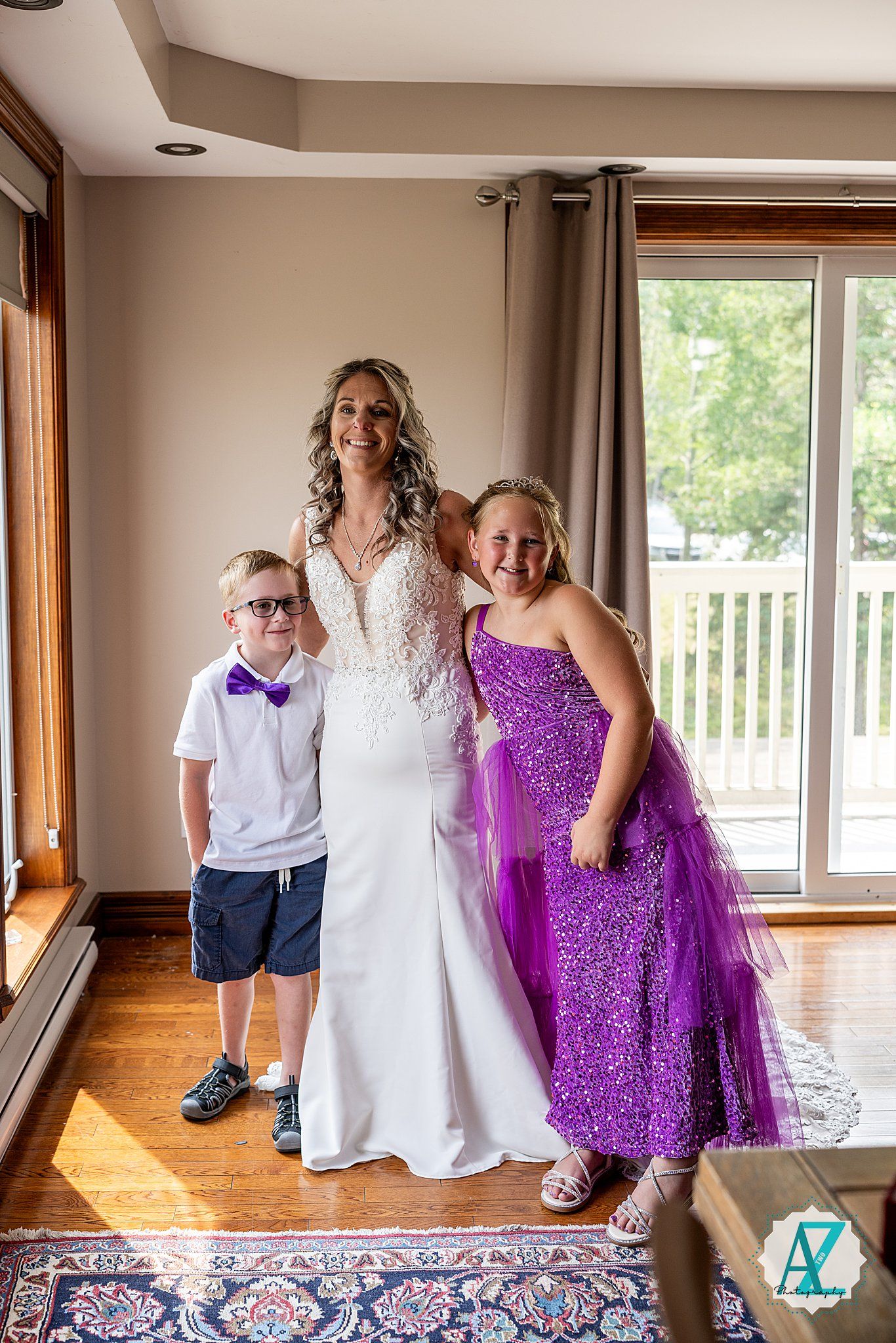 Wedding group photo in home interior with white wedding gown and bright purple dress against hardwood floor and window.